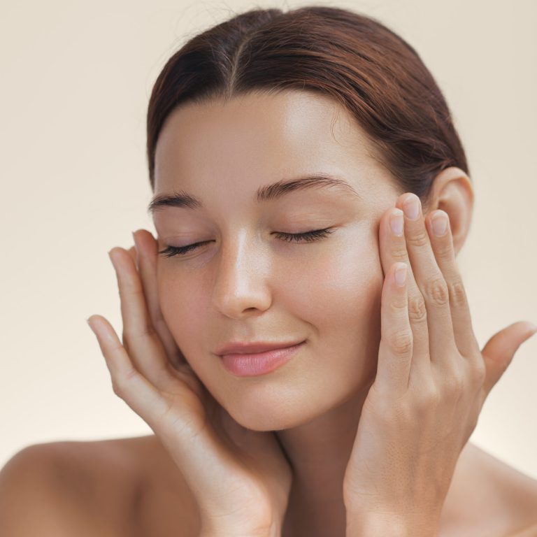 Cosmetics skincare concept photo. Woman with beautiful face touching healthy facial skin portrait. Beautiful smiling Asian girl model with natural makeup enjoys glowing hydrated smooth skin on beige background closeup. High-quality photo